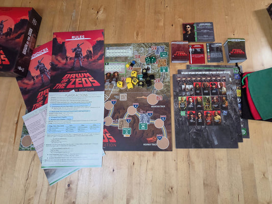 A tabletop layout of the board game 'Blind Date with the Zeds' showing various components including the main board, instruction booklet, cards, tokens, character sheets, a green drawstring pouch, and boxes on a light wood floor.