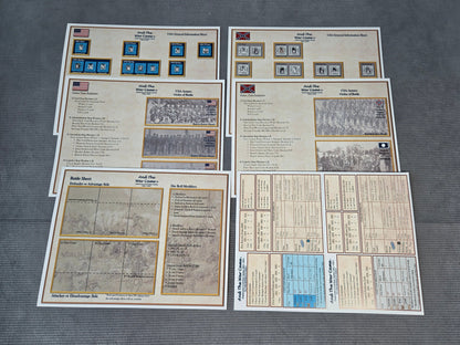 Four printed game boards or playmat sheets laid out on a dark gray textured surface. The boards feature beige backgrounds with printed maps, rules text, grid sections, and small blue and red icon tiles.