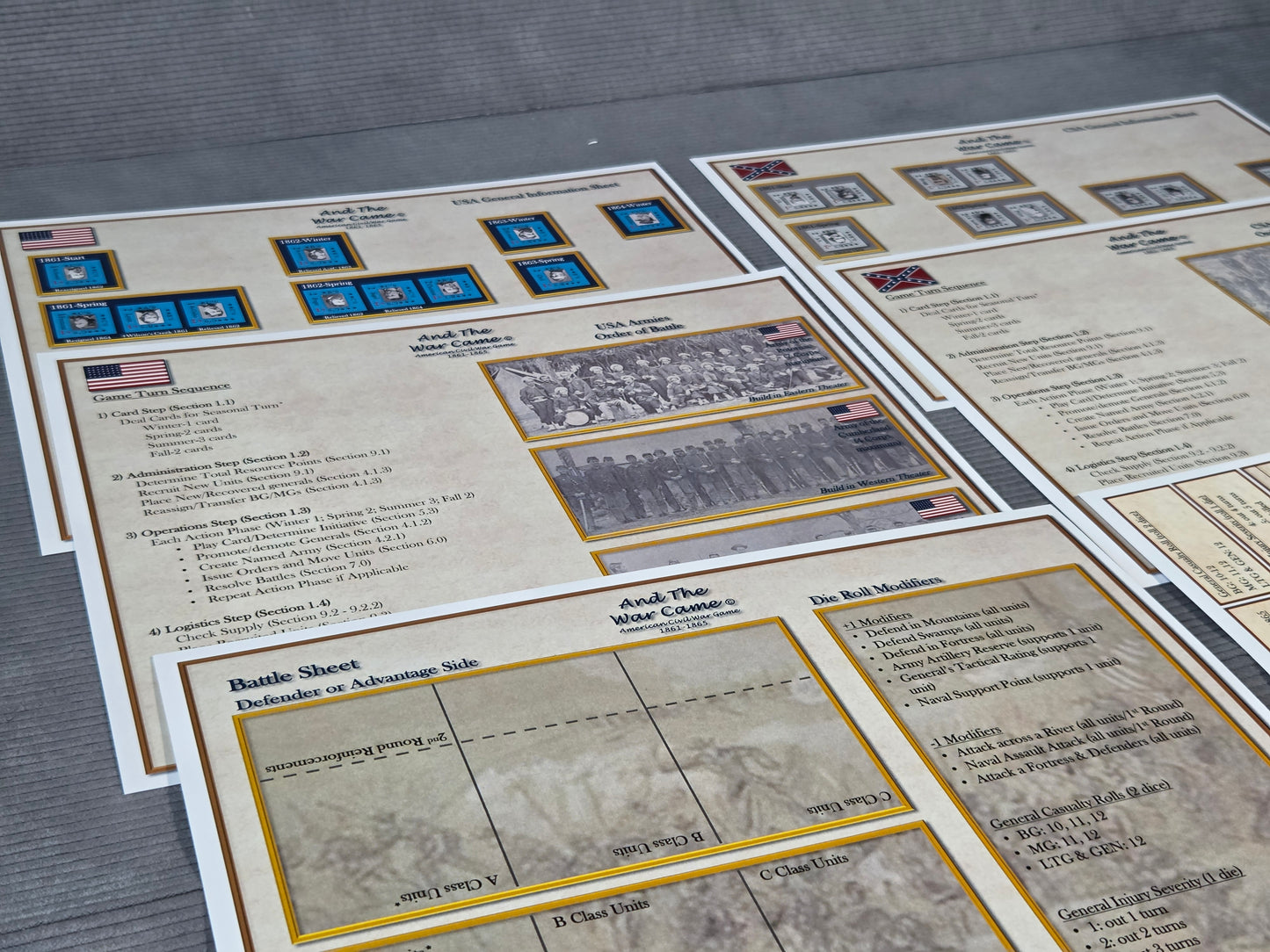 Several printed historical battle sheets and informational cards arranged on a gray surface. The cards have aged beige paper, gold border lines, small blue insignia, black text, and some black-and-white historical photographs.