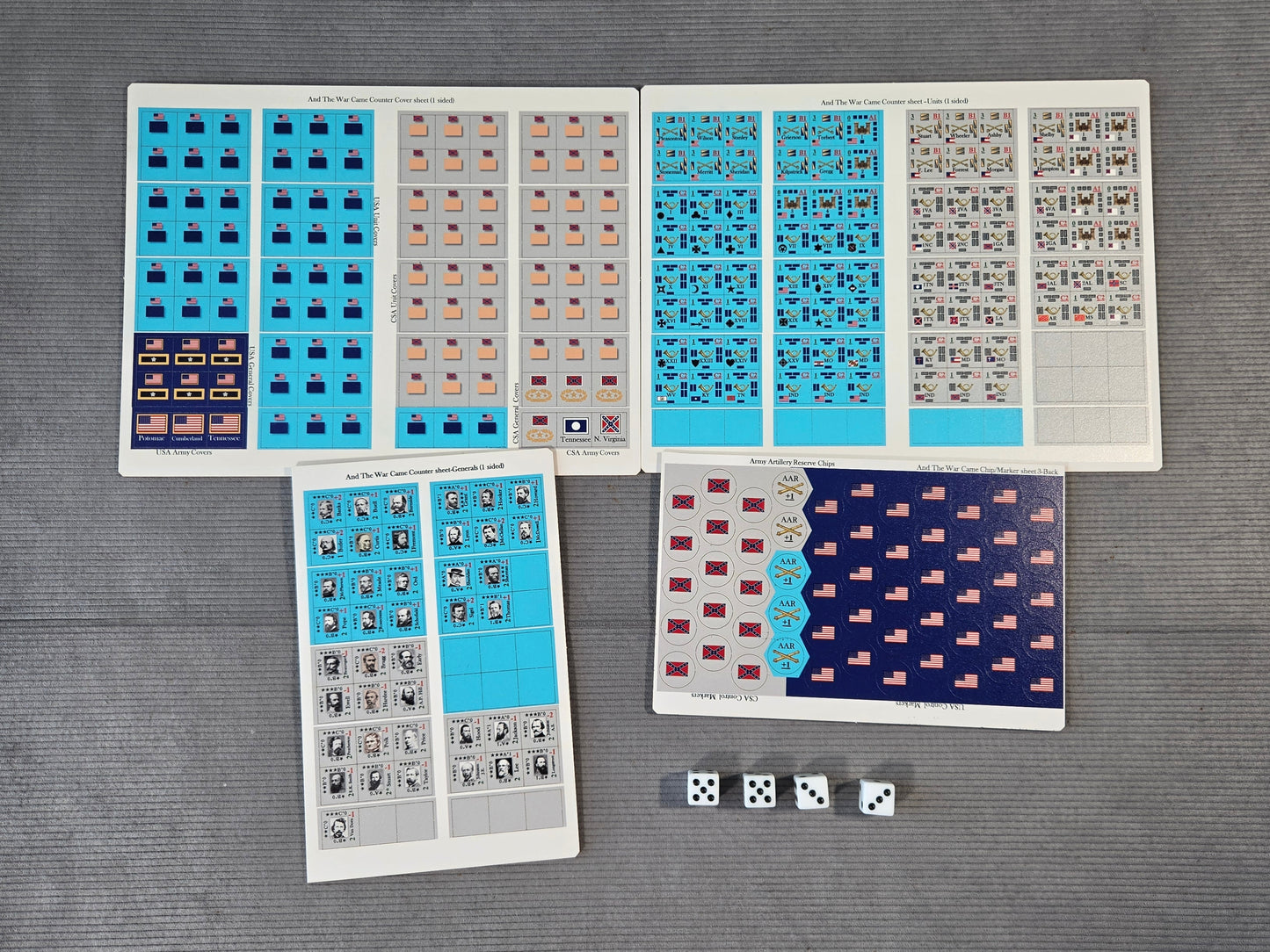A set of printed game or activity sheets laid out on a gray textured surface, including blue, beige, gray, and dark navy patterned cards or stickers, plus three white dice.
