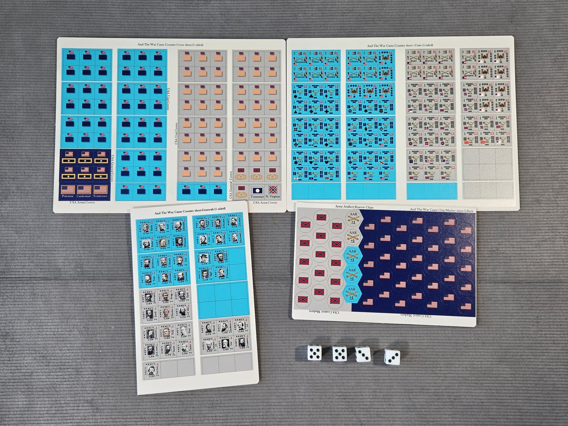 A set of printed game or activity sheets laid out on a gray textured surface, including blue, beige, gray, and dark navy patterned cards or stickers, plus three white dice.