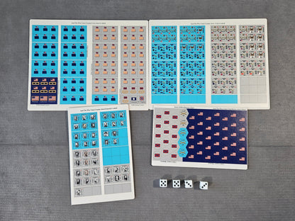 A set of printed game or activity sheets laid out on a gray textured surface, including blue, beige, gray, and dark navy patterned cards or stickers, plus three white dice.