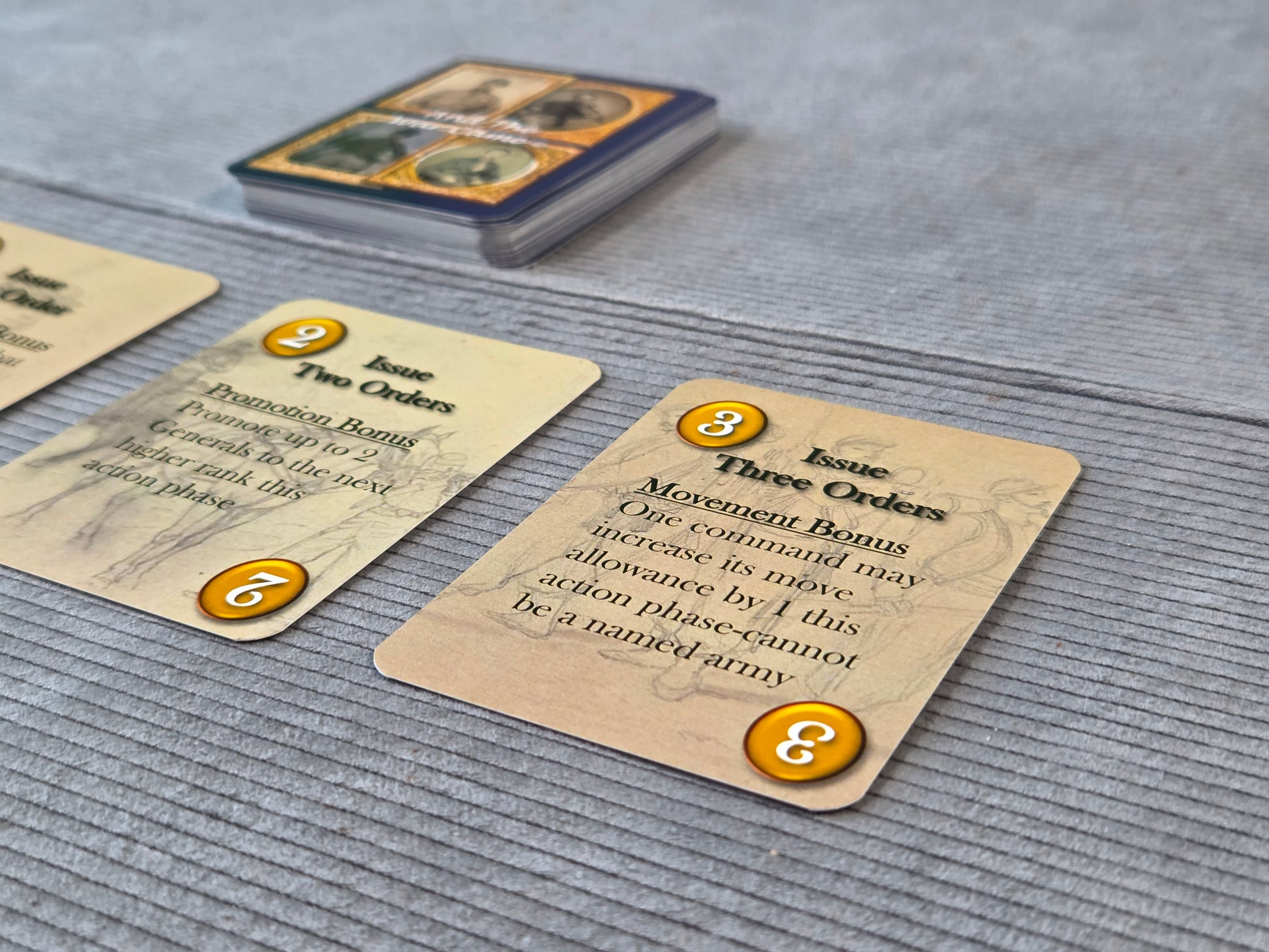 Close-up of tabletop gaming cards on a gray fabric surface, with a deck of cards visible in the background. The cards are tan with black text and orange circular number badges.
