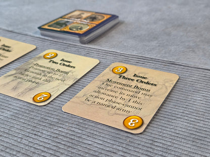 Close-up of tabletop gaming cards on a gray fabric surface, with a deck of cards visible in the background. The cards are tan with black text and orange circular number badges.