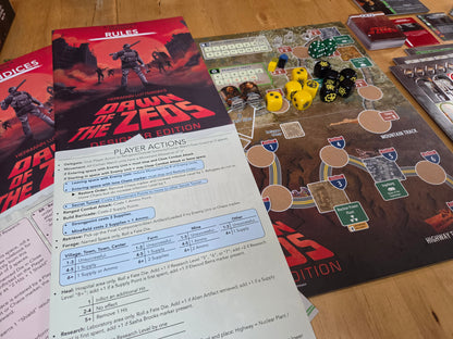 A tabletop view of a board game setup showing the 'Drawn of the Zeds' Player Actions sheet, game box cover, playmat, dice, and cards on a wooden surface.