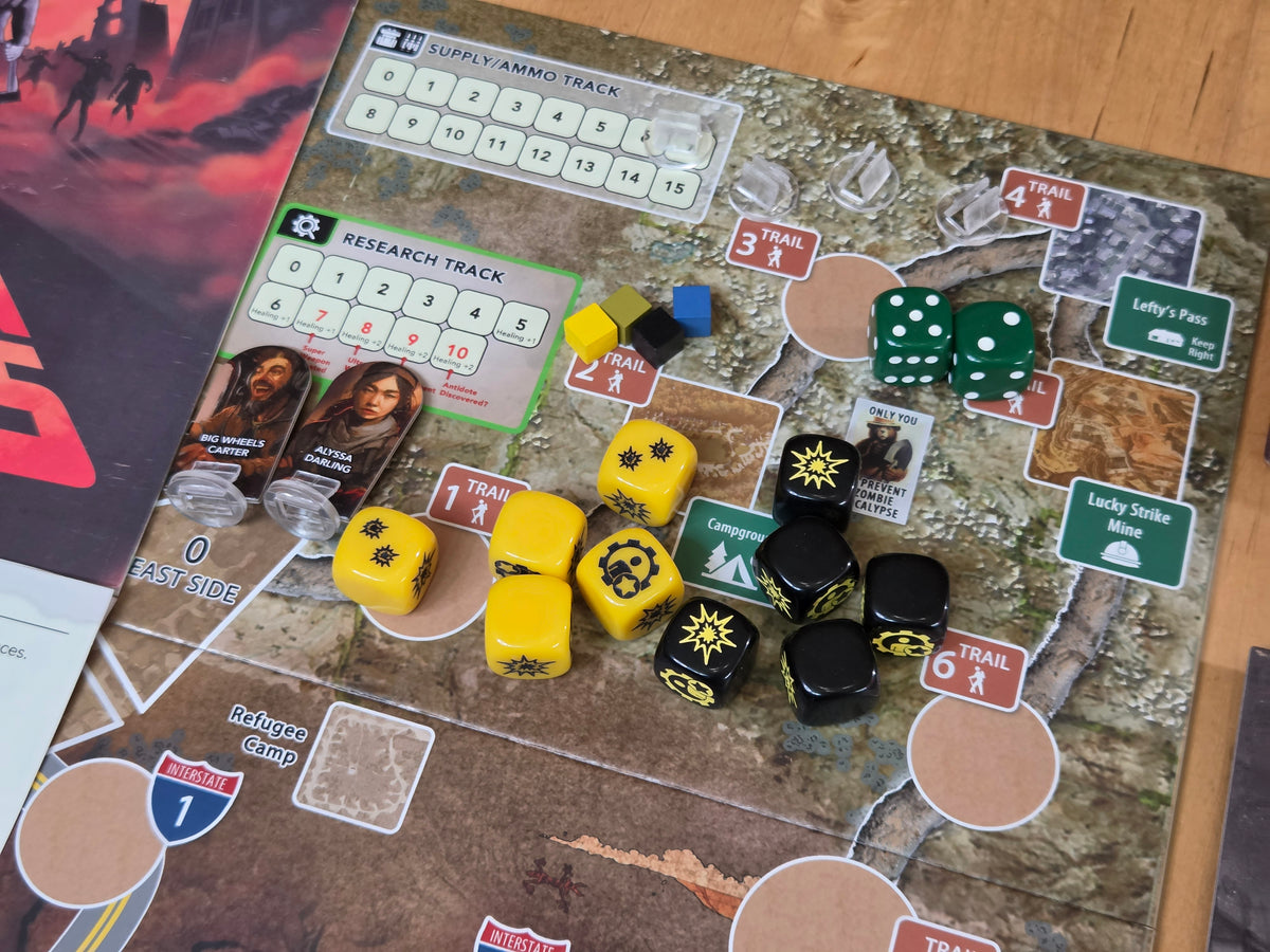 A close-up of a board game setup on a map-style playmat, showing yellow and black dice, green dice, cards, and small game pieces on a wooden tabletop.