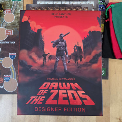 The board game box for 'Dawn of the Zeeds Designer Edition' by Blue Panther is displayed on a light wood tabletop, surrounded by game components including a playmat, cards, dice, and green/red draw bags.