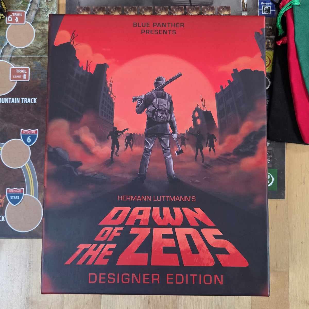 The board game box for 'Dawn of the Zeeds Designer Edition' by Blue Panther is displayed on a light wood tabletop, surrounded by game components including a playmat, cards, dice, and green/red draw bags.
