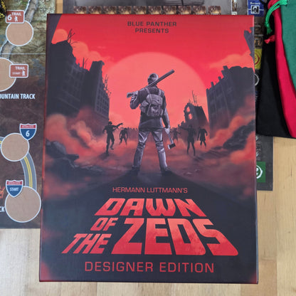 The board game box for 'Dawn of the Zeeds Designer Edition' by Blue Panther is displayed on a light wood tabletop, surrounded by game components including a playmat, cards, dice, and green/red draw bags.